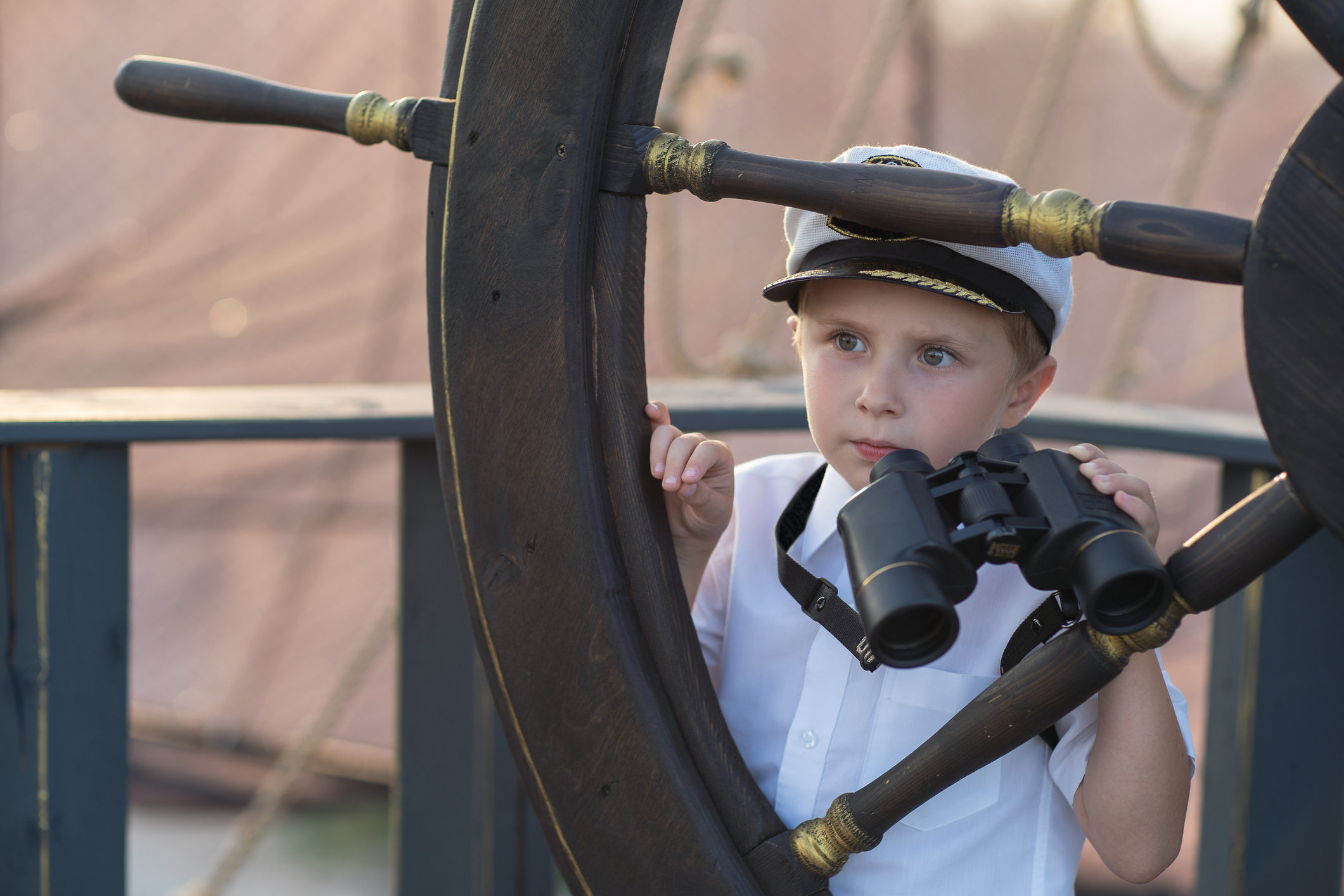 Little boy playing ship captain Little ship captain on the deck of a pirate ship. The boy stands on the deck of the ship behind the marine steering wheel. The boy is holding marine binoculars. The boy looks into the distance.