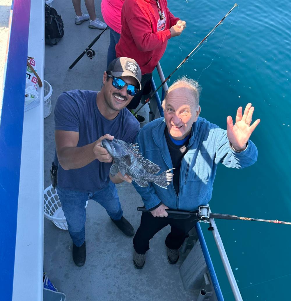 Two men on a boat with a fish; one holding it, the other smiling and waving.