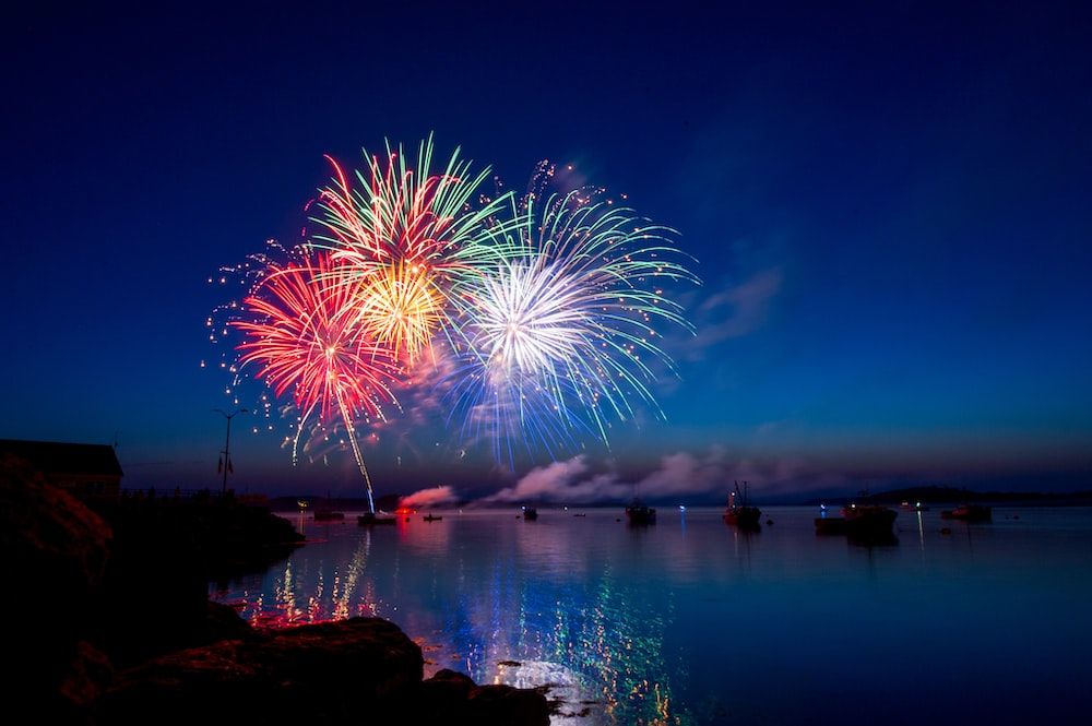 Colorful fireworks over calm water at dusk, with reflections on the surface.
