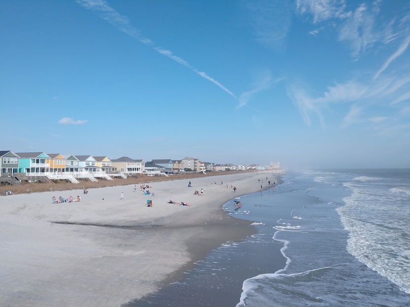 Beachfront with colorful houses, people relaxing, and gentle waves under a blue sky.