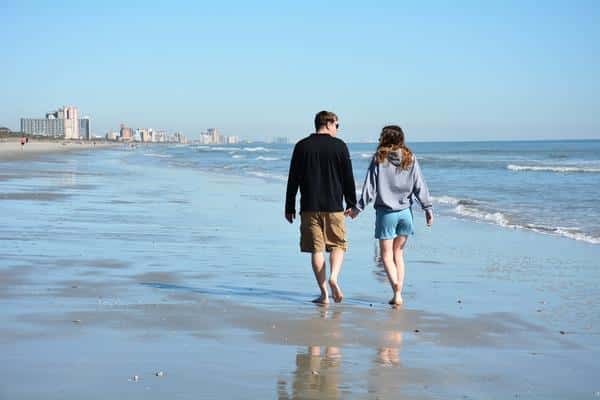 Couple walking barefoot on a sandy beach with city skyline in the distance.